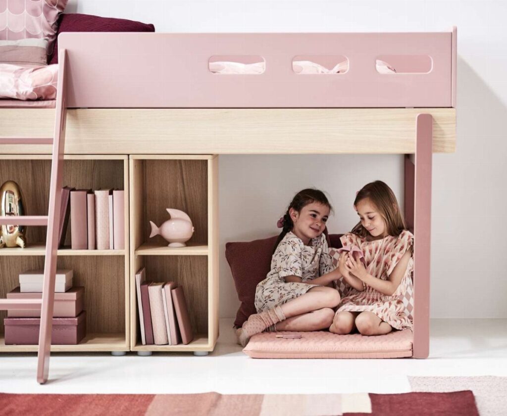 Two girls sitting under a pink and wood loft bed with safety rails.