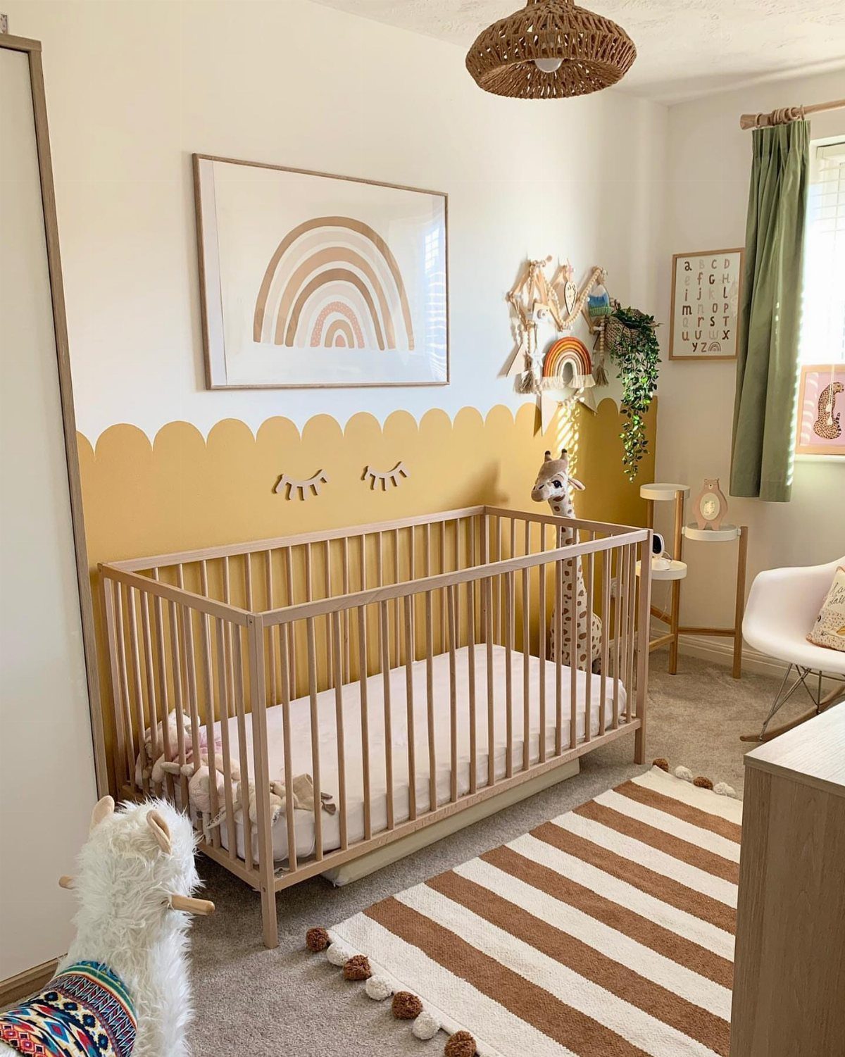 Boho nursery with a light wood crib, rainbow art, and striped pom-pom rug.