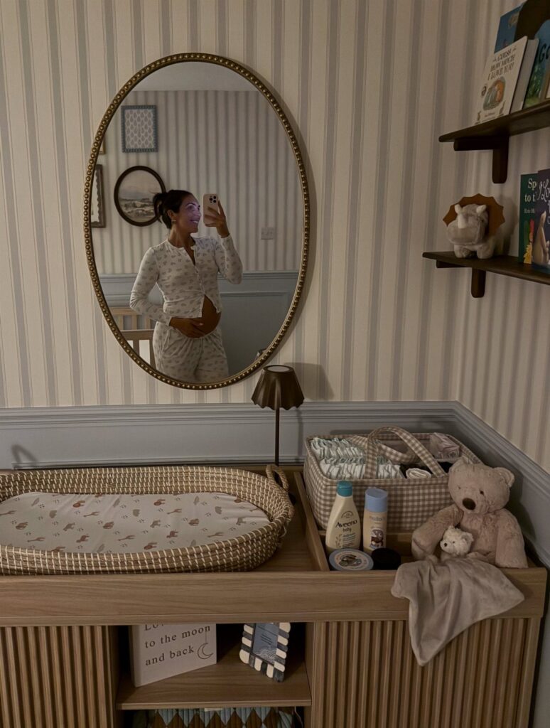 Pregnant woman in a nursery with a wooden changing table and striped wallpaper.