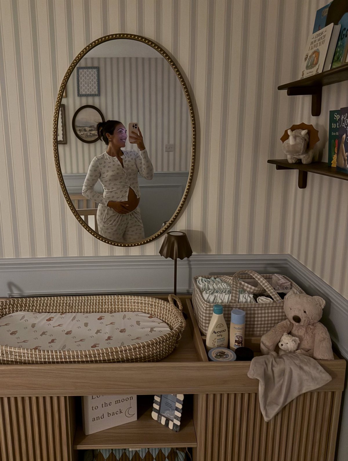 Pregnant woman in a nursery with a wooden changing table and striped wallpaper.