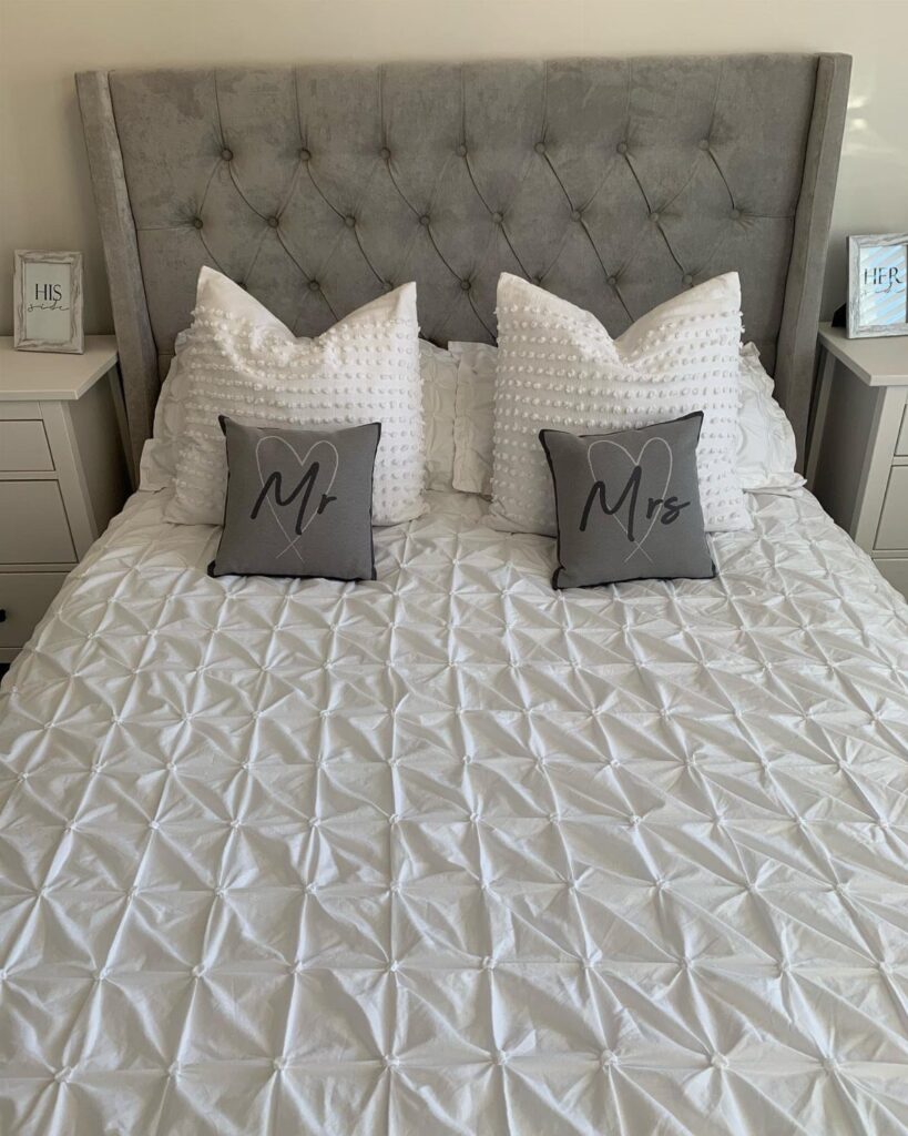 Bed with grey tufted headboard, white textured linens, and Mr and Mrs pillows.