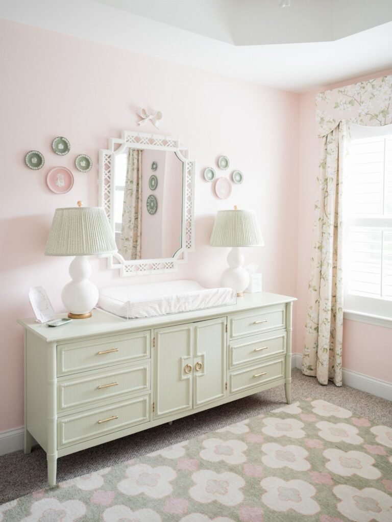 Pink and green nursery with white dresser, floral rug, and patterned lampshades.
