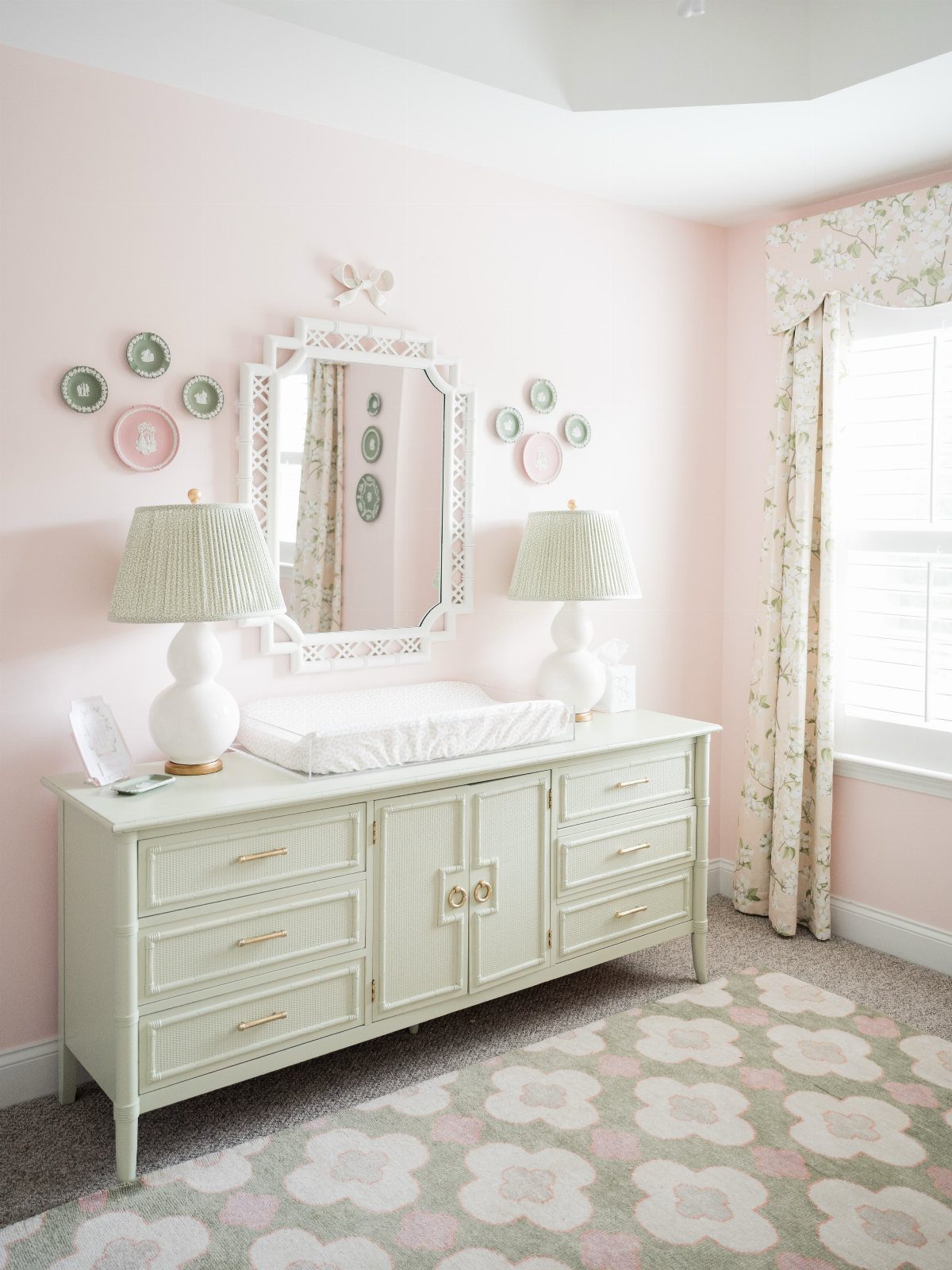 Pink and green nursery with white dresser, floral rug, and patterned lampshades.