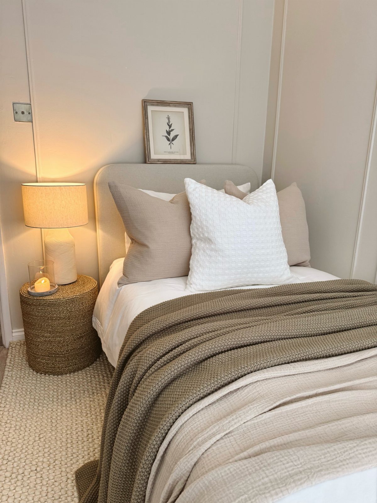 Small bedroom with neutral bedding, a woven side table, and a warm lamp.